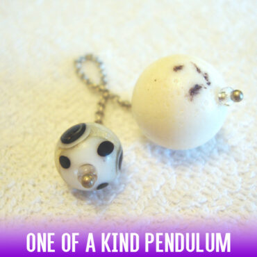 A dowsing pendulum made of a white, natural sandstone round bead with black details, a silver chain and a black-white-gold handle