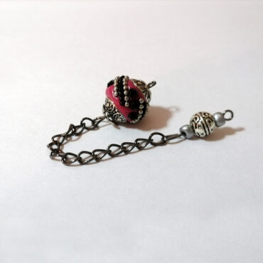 A dowsing pendulum made with a handmade antique silver alloy deep magenta Indonesian bead with black rhinestones crystals and tiny silver embedded beads. There are vintage style embedded end caps and a gun colored antique chain. The holder is made with a petite metallic alloy handcrafted bead. A very Asian exotic feel.