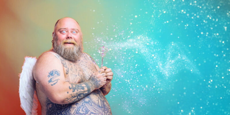 Spiritual Narcissism: A bearded, topless and heavy-set man with tattoos and wings looks amazed and acts like a fairy who is sprinkling fairy dust